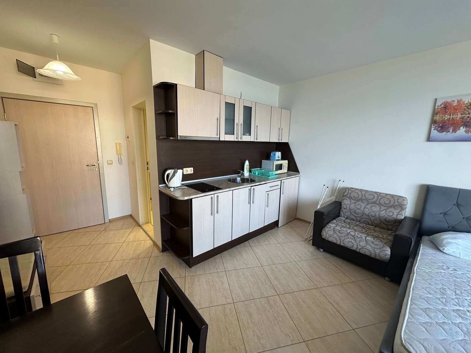 Современная студия с кухней, диваном и обеденным столом; светлые тона, плитка на полу, окно с естественным освещением.