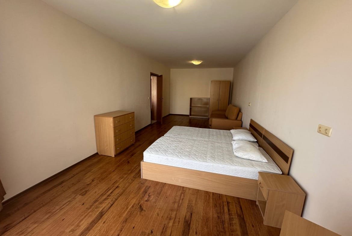 Спальня с деревянным полом, двуспальная кровать, комод и диван. Светлые стены и минималистичный интерьер.