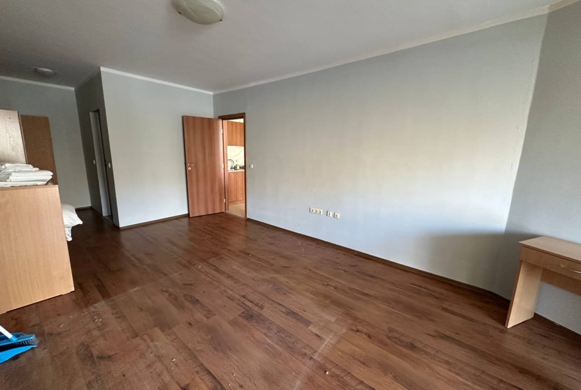 Пустая комната с деревянным полом, двумя дверями и мебелью, включая стол и шкаф с постельным бельем.