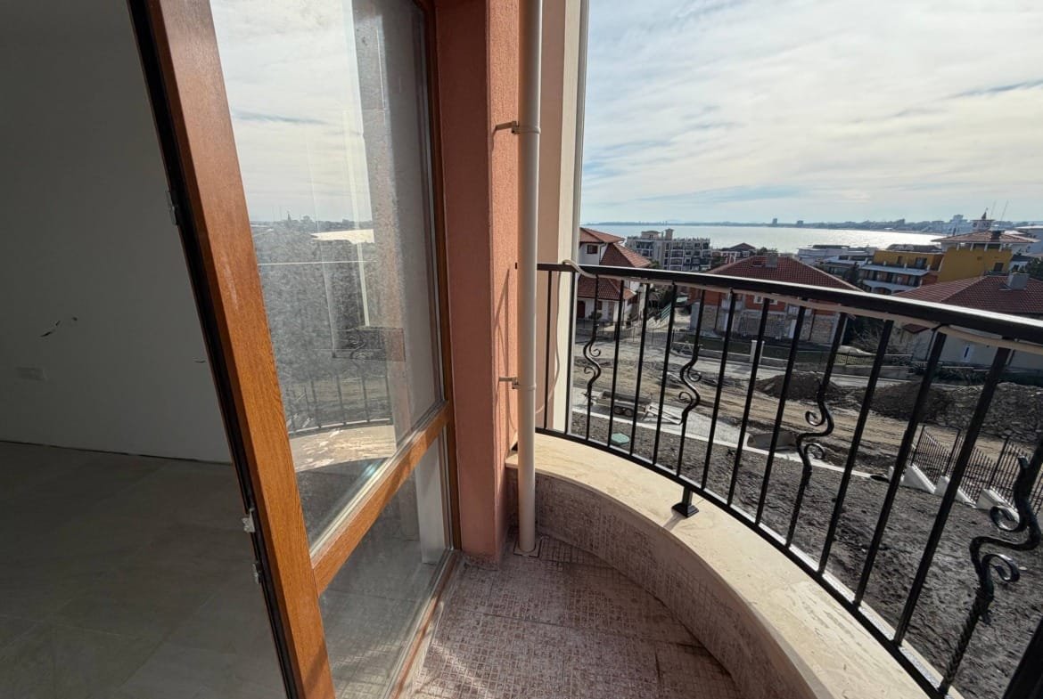 Балкон с деревянной дверью, открывающейся на панорамный вид, море и жилые дома на фоне ясного неба.