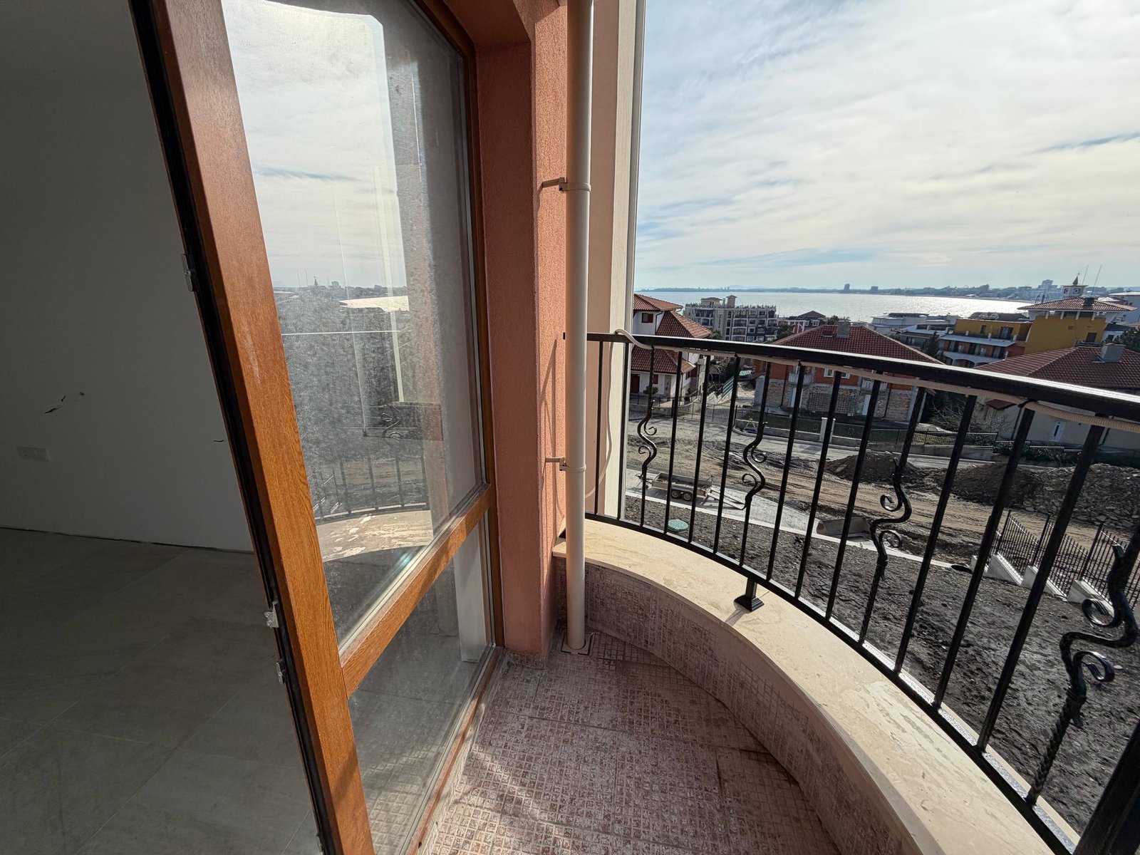 Балкон с деревянной дверью, открывающейся на панорамный вид, море и жилые дома на фоне ясного неба.