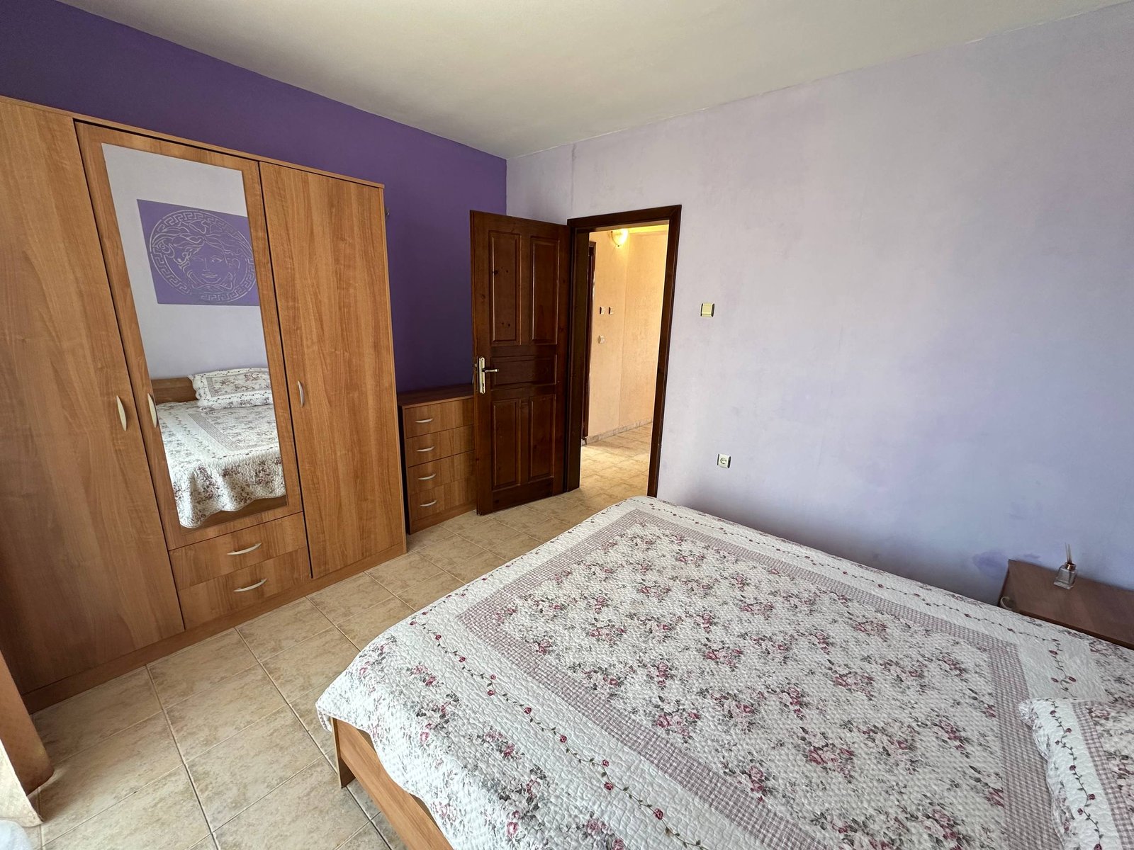 Комната с двуспальной кроватью, комодом и зеркальным шкафом, стены окрашены в светло-фиолетовый цвет.