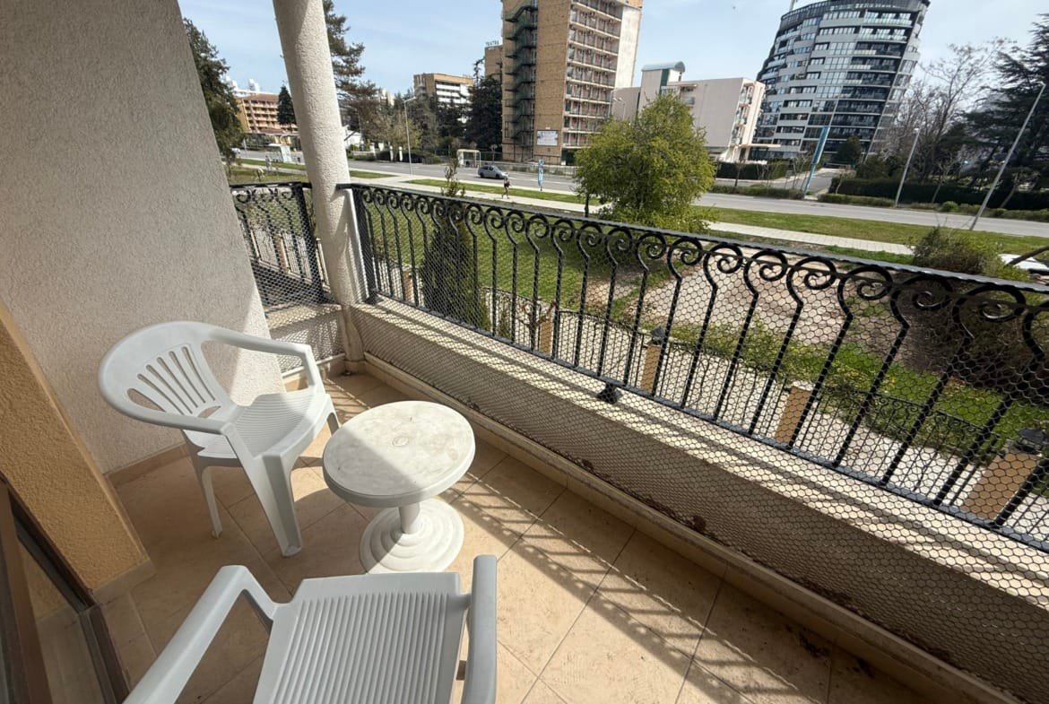 Балкон с двумя пластиковыми креслами и столом, вид на застройку и зеленую территорию, идеальное место для отдыха в Болгарии.