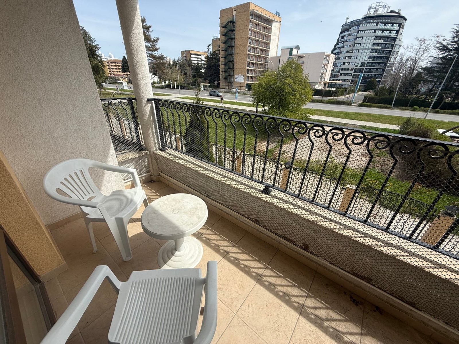 Балкон с двумя пластиковыми креслами и столом, вид на застройку и зеленую территорию, идеальное место для отдыха в Болгарии.