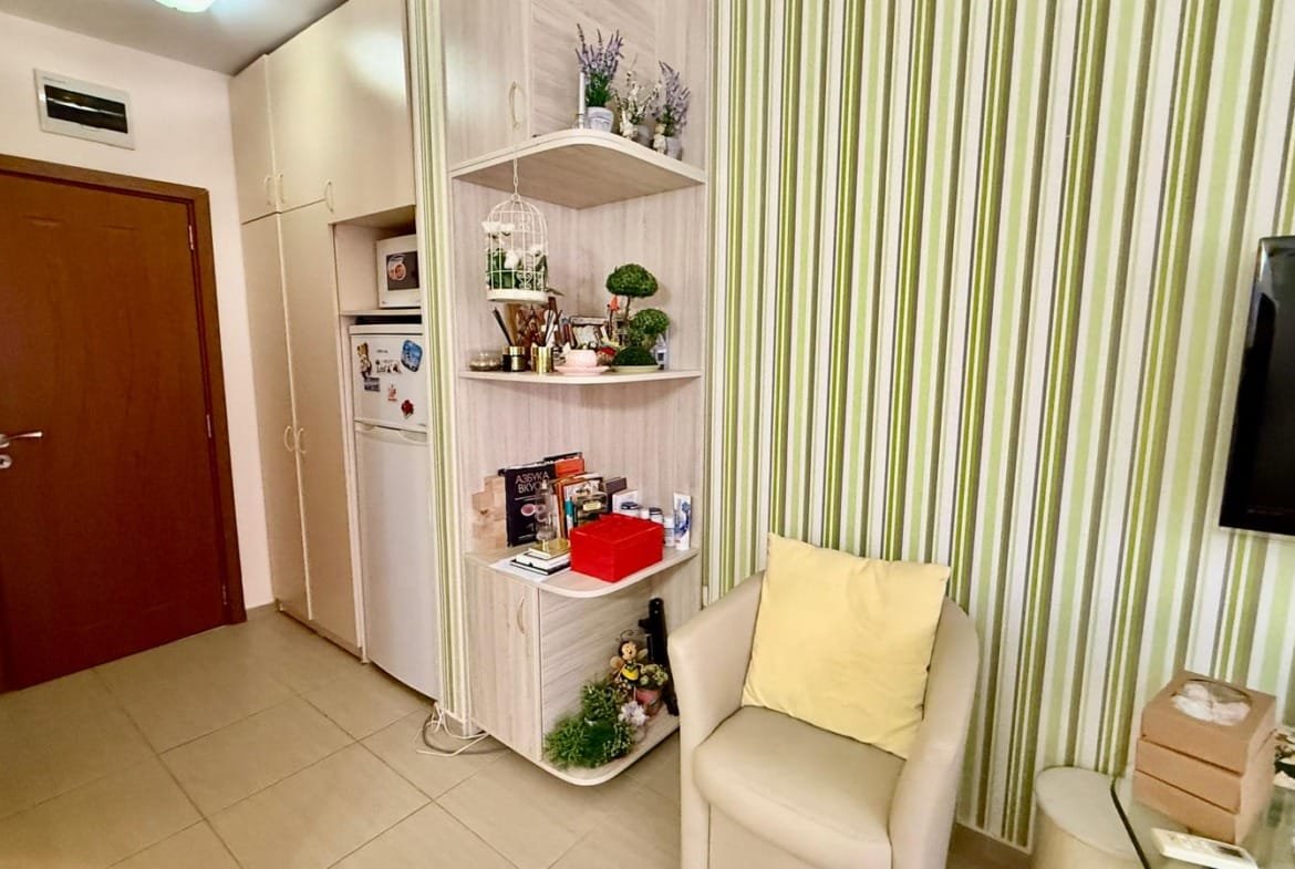 Уютный уголок квартиры с полкой для декора, мягким креслом и зелеными стенами, подходящий для покупки недвижимости в Болгарии.