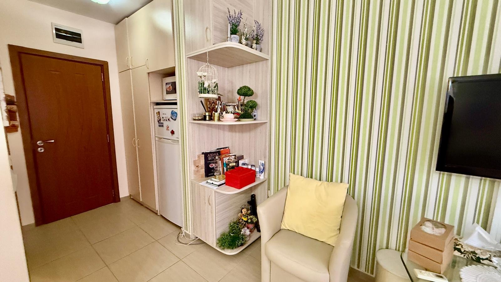 Уютный уголок квартиры с полкой для декора, мягким креслом и зелеными стенами, подходящий для покупки недвижимости в Болгарии.