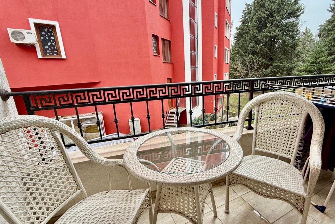 Балкон с белыми плетеными креслами и столом, зелеными деревьями и красным зданием на фоне, идеальный для отдыха в Болгарии.
