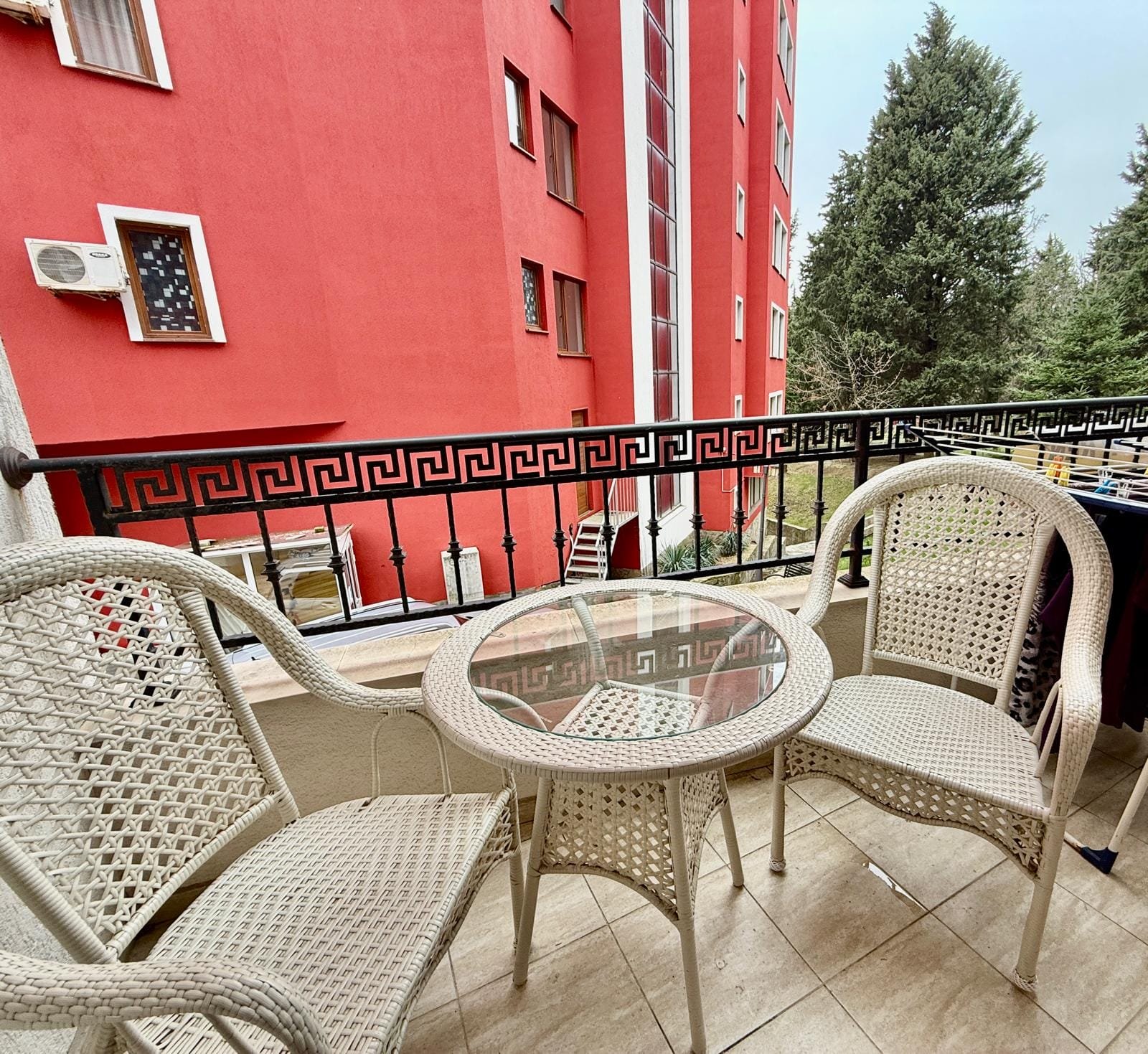 Балкон с белыми плетеными креслами и столом, зелеными деревьями и красным зданием на фоне, идеальный для отдыха в Болгарии.