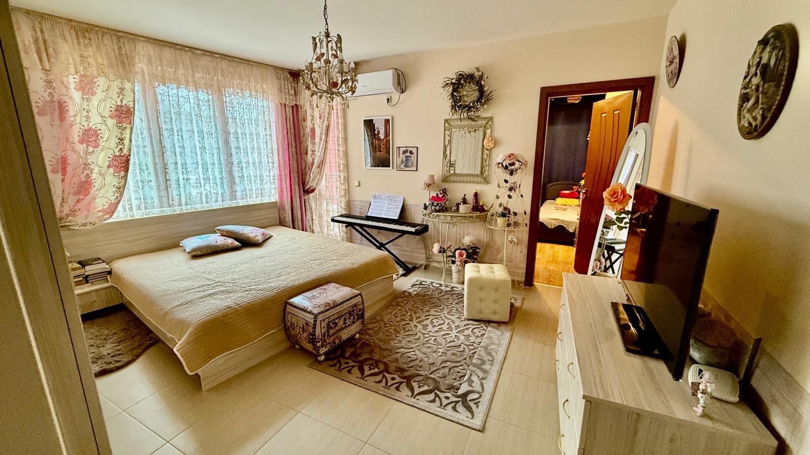 Современная спальня с двуспальной кроватью, красивыми шторами и телевизором, стильный интерьер для комфортной жизни в Болгарии.