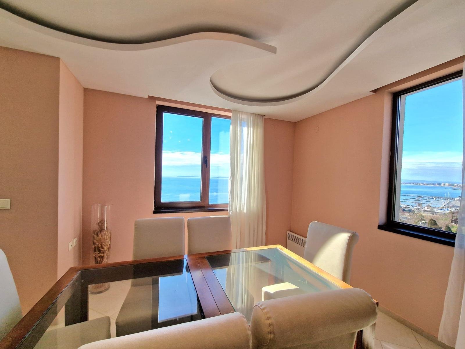 Светлая гостиная с панорамным окном, столом и стульями, предлагающая вид на море в Болгарии. Идеальное место для отдыха.