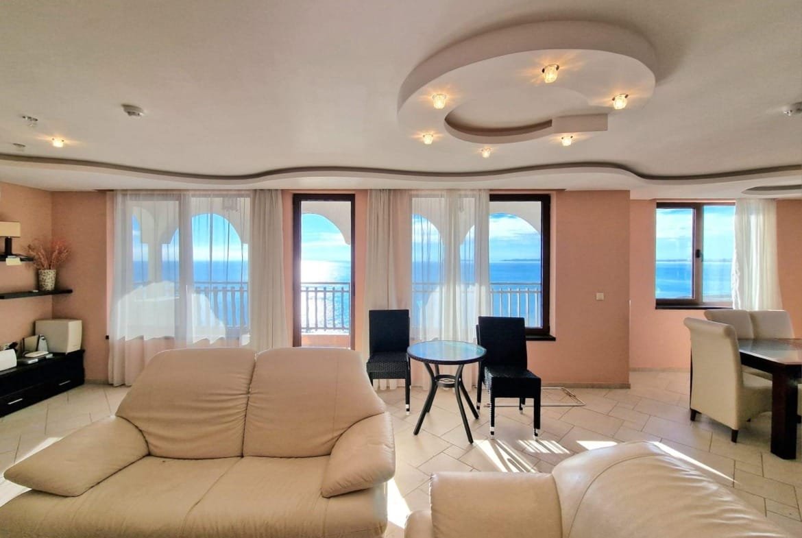 Современная гостиная с панорамным видом на море, светлые стены и уютная мебель, идеальное место для жизни в Болгарии.