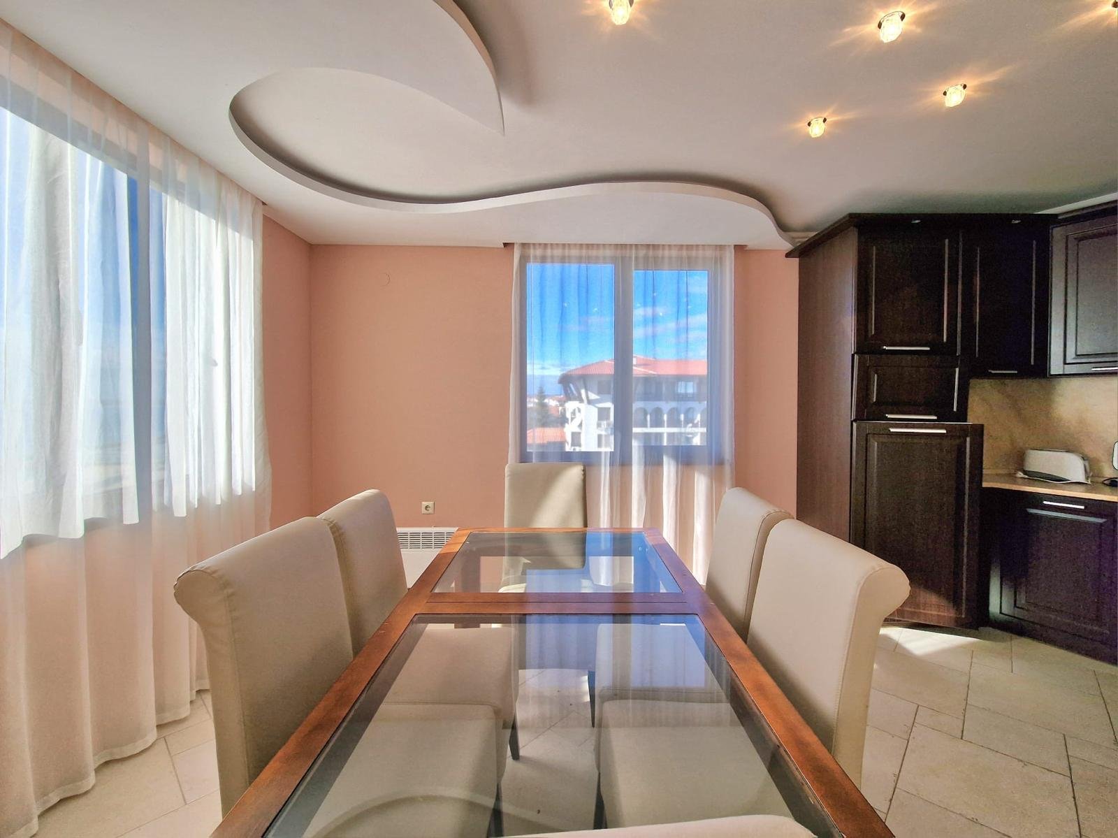 Современный интерьер квартиры в Болгарии, светлое помещение с окнами, стеклянным столом и уютной мебелью.