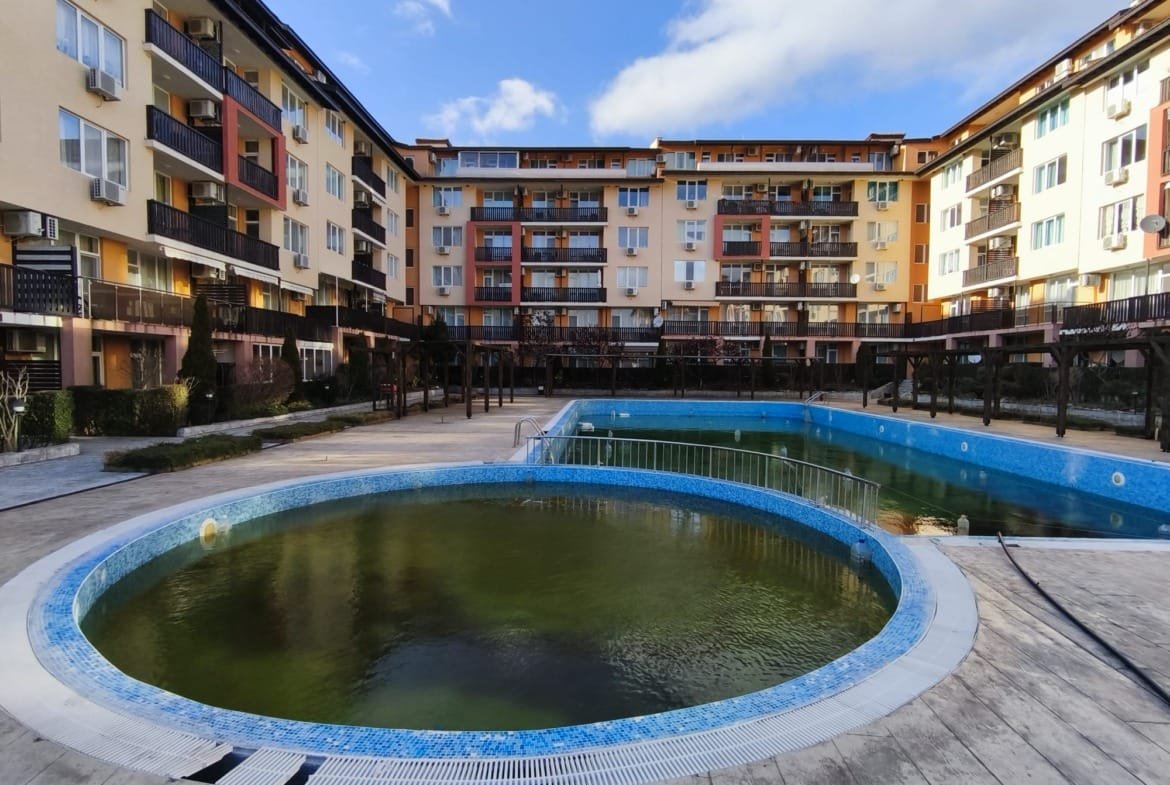 Комплекс жилой недвижимости в Болгарии с бассейном, окружённый зданиями. Яркое небо и зелёные растения создают уютную атмосферу.