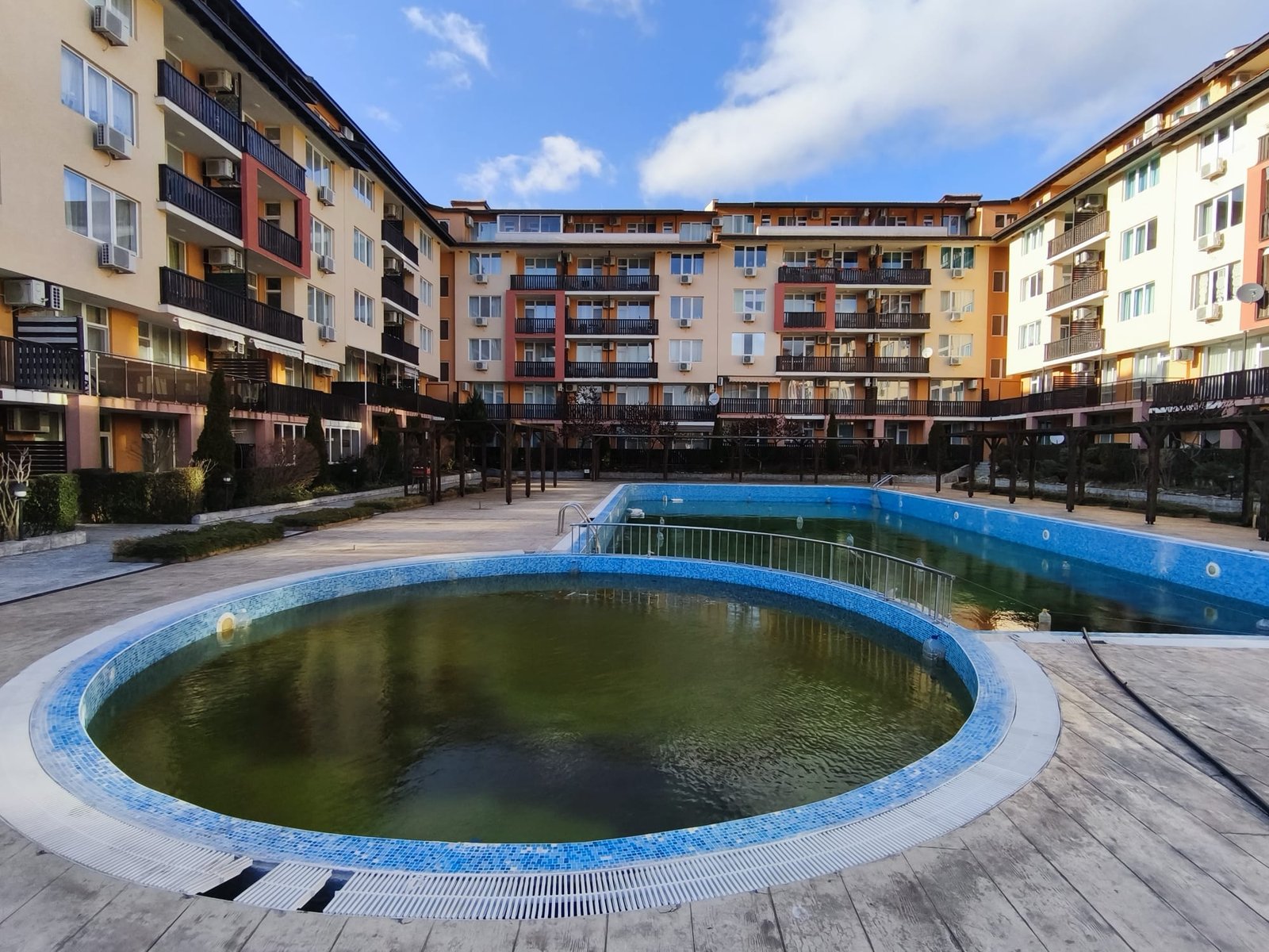 Пустой бассейн в жилом комплексе с многоквартирными домами Комплекс жилой недвижимости в Болгарии с бассейном, окружённый зданиями. Яркое небо и зелёные растения создают уютную атмосферу.