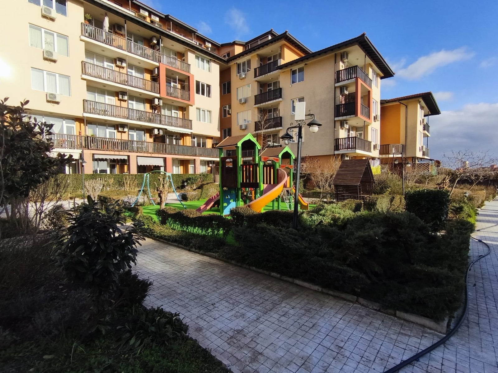 Детская площадка во дворе жилого комплекса на фоне зданий Жилой комплекс в Болгарии с площадкой для детей, зелеными насаждениями и ухоженной территорией для комфортного проживания.