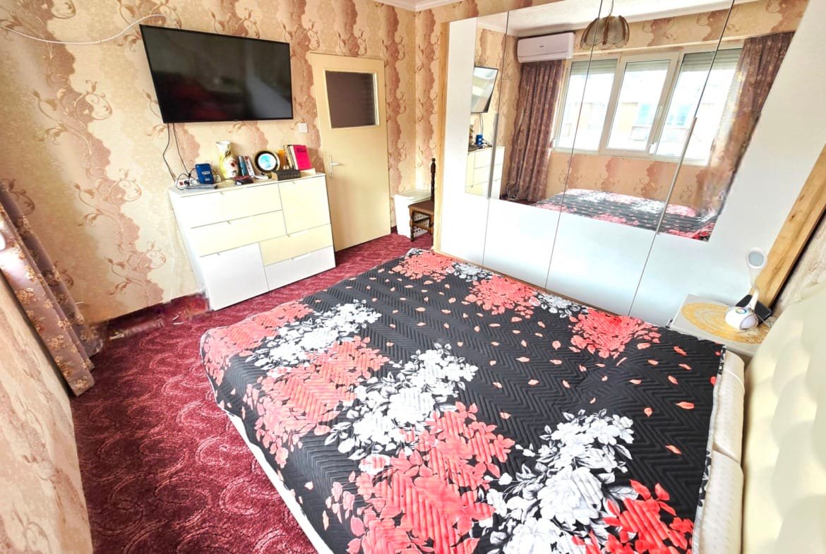 Уютная спальня с двуспальной кроватью, телевизором, шкафом с зеркальными дверями и ярким текстилем для продажи недвижимости в Болгарии.