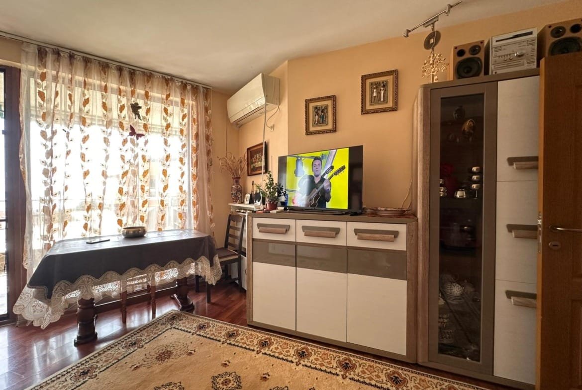 Уютная гостиная с телевизором, столом и шкафом, оформленная в теплых тонах, подходит для жизни и аренды в Болгарии.