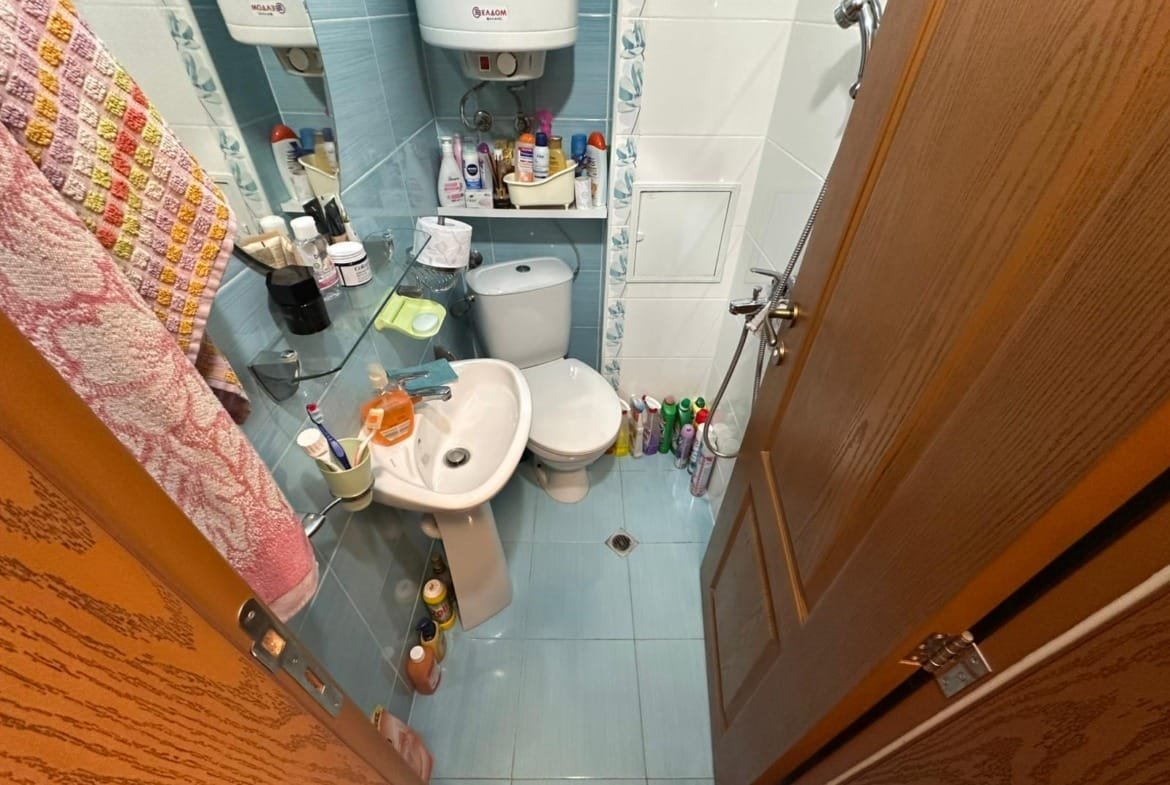Маленькая ванная комната с сантехникой, зеркалом и полками для косметики, оформленная в светлых тонах, подходит для покупки недвижимости в Болгарии.