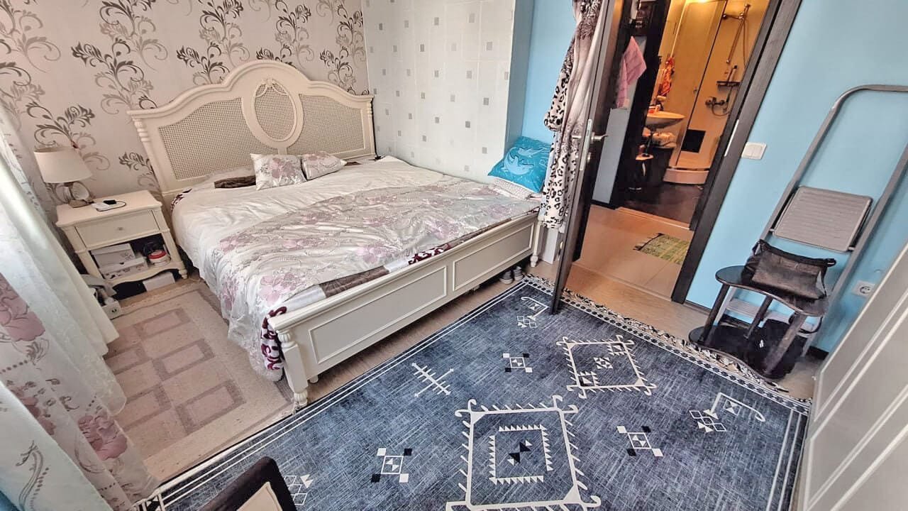 Уютная спальня с двуспальной кроватью, окрашенными в светлые тона стенами и ванной в светлом углу, идеально для покупки недвижимости в Болгарии.
